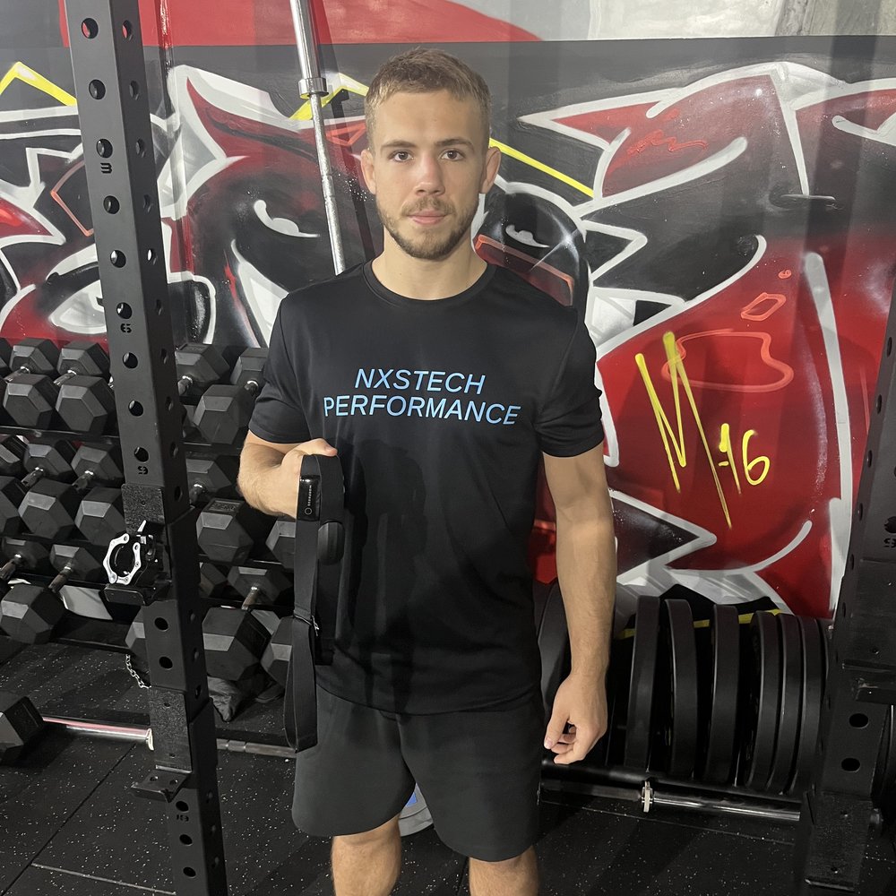Krane in an NxsTech Performance shirt holding a heart rate strap, M-16 graffiti on the wall behind him.