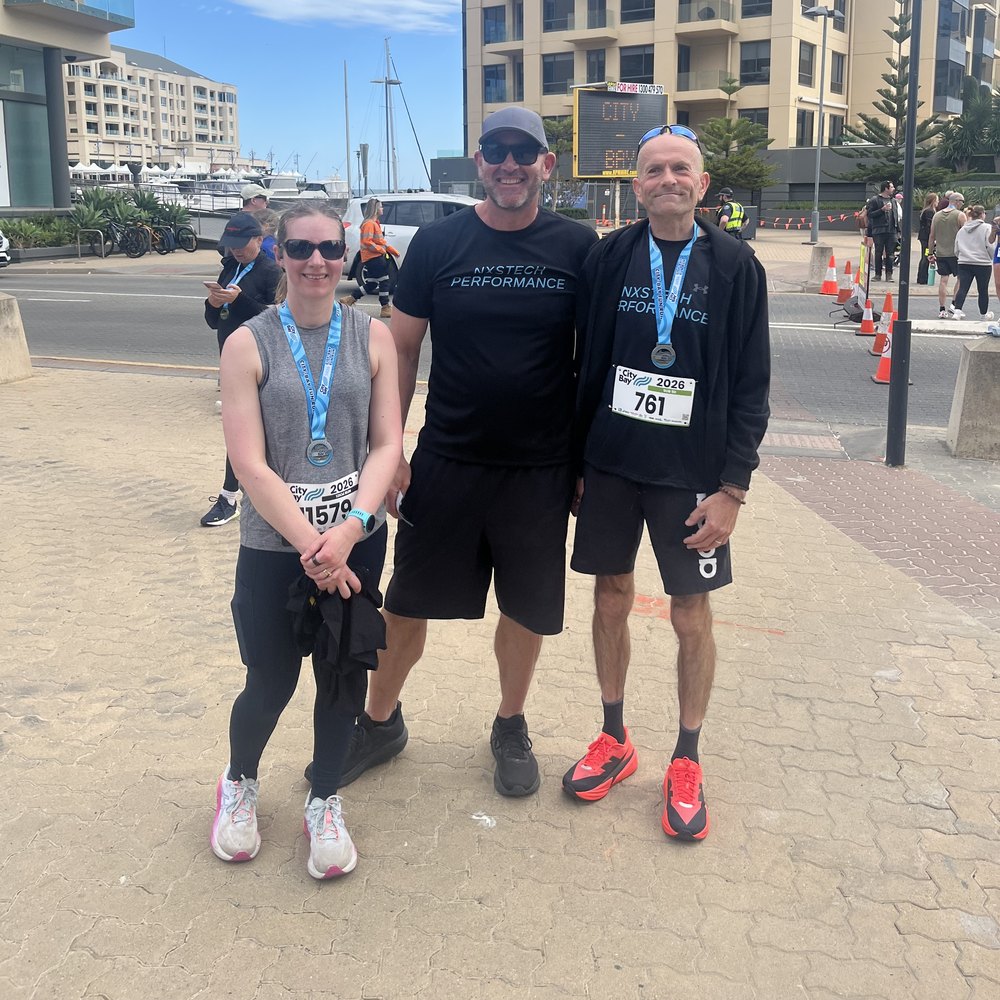 Three runners standing together after a race, wearing City-Bay 2026 bibs and finisher medals, two of them in NxsTech Performance shirts.